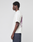 A person with short curly hair, wearing the LIVE FAST DIE YOUNG LFDY Grand Theft Tee—an oversized white 100% Baumwolle T-shirt with back text and graphics—and dark jeans stands in profile against a plain light gray background.