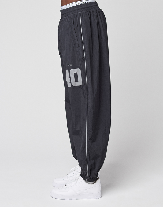 A person is shown in profile wearing LIVE FAST DIE YOUNG’s LFDY Performance Trackpants—loose-fitting, black, with a reflective print and the number 40, styled in an oversize fit with white sneakers against a plain white background.