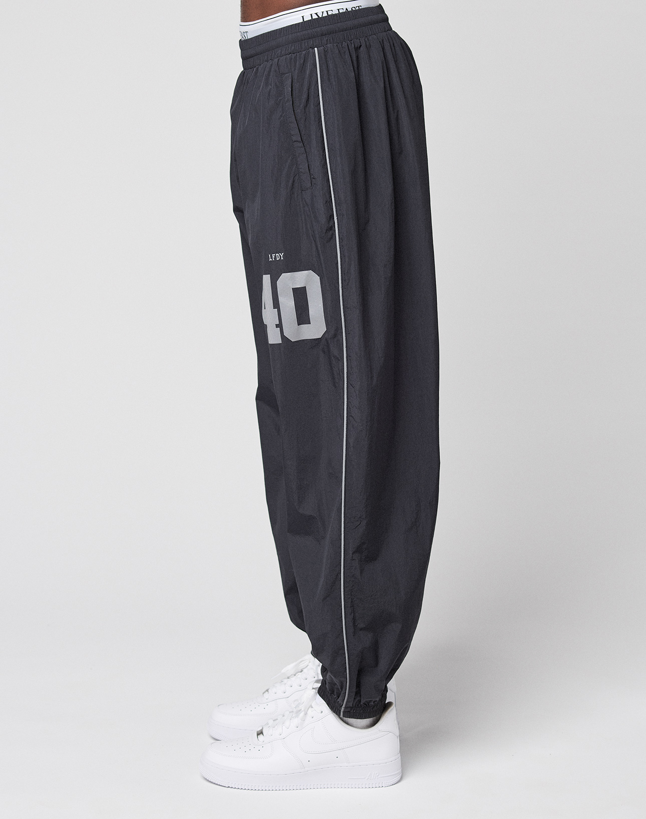 A person is shown in profile wearing LIVE FAST DIE YOUNG’s LFDY Performance Trackpants—loose-fitting, black, with a reflective print and the number 40, styled in an oversize fit with white sneakers against a plain white background.