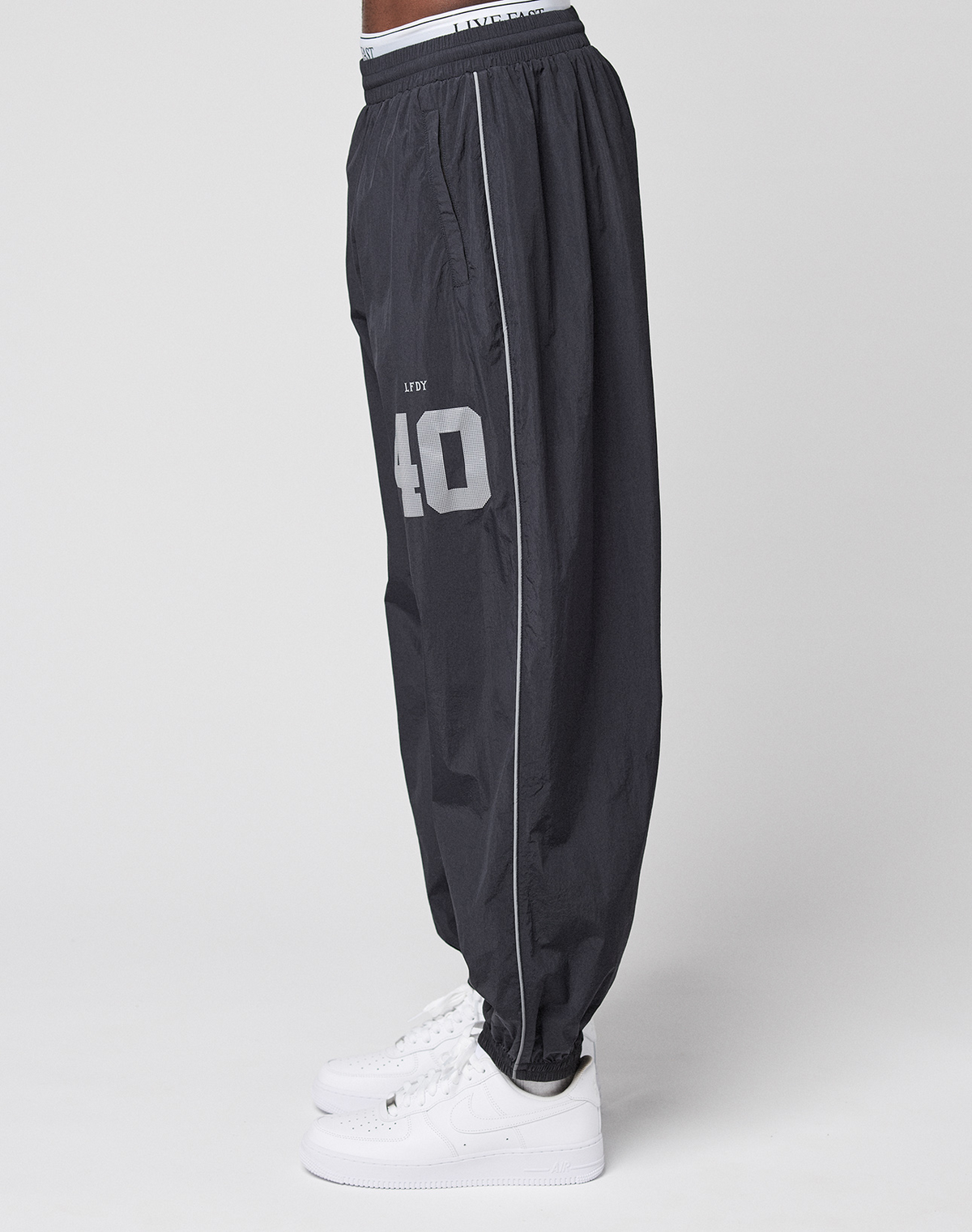 A person is shown in profile wearing LIVE FAST DIE YOUNG’s LFDY Performance Trackpants—loose-fitting, black, with a reflective print and the number 40, styled in an oversize fit with white sneakers against a plain white background.