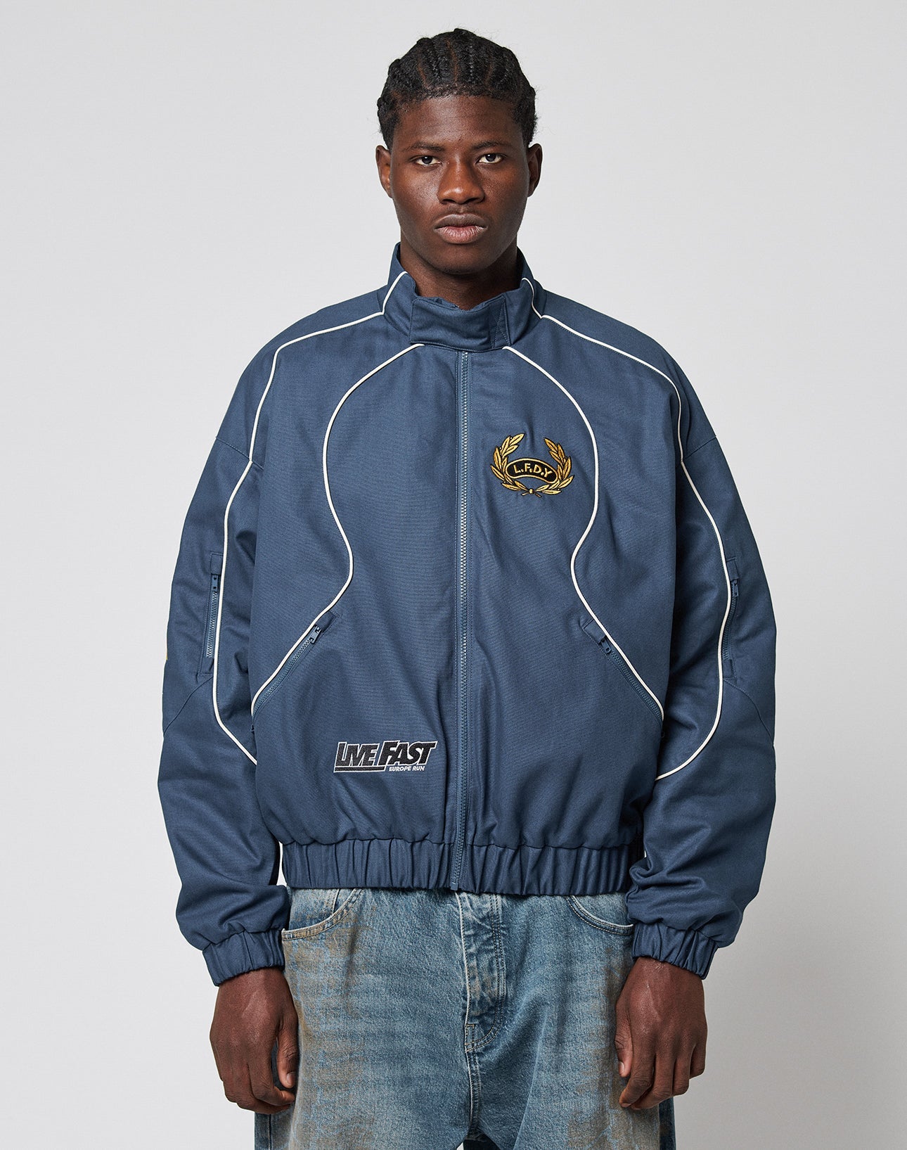 A man wearing an LFDY Canvas Jacket by LIVE FAST DIE YOUNG with white piping, multi-logo embroidery, elastic cuffs, and baggy blue jeans stands against a simple light background, looking forward.