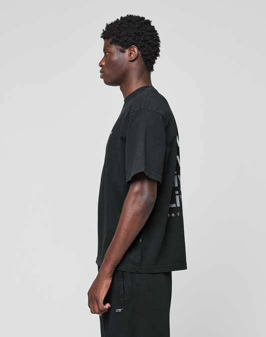 A person with short curly hair stands in profile against a plain background, wearing the oversize fit Basic Evidence Tee by LIVE FAST DIE YOUNG, made of 100% Baumwolle, paired with black pants.