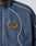Close-up of the LFDY Canvas Jacket by LIVE FAST DIE YOUNG, featuring elastic cuffs, blue fabric, white piping, and an embroidered yellow laurel wreath with L.F.D.Y letters stitched in yellow on a black background.