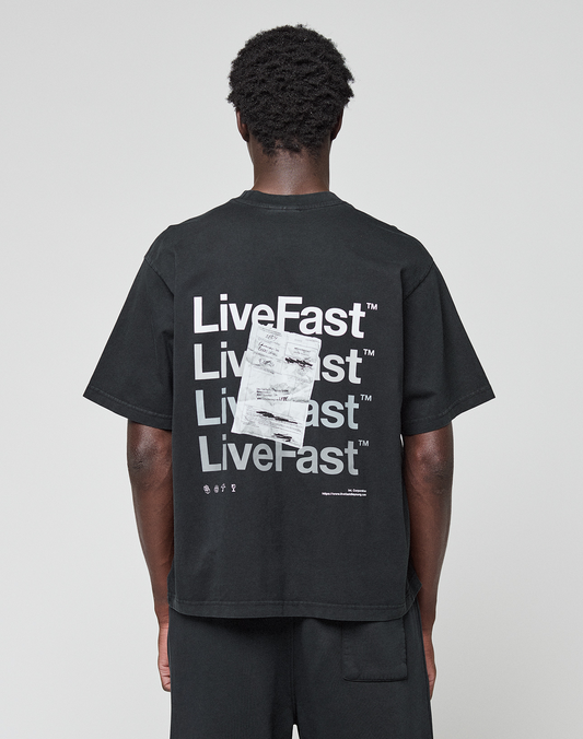 A person, facing away from the camera, wears the LIVE FAST DIE YOUNG Basic Evidence Tee—an oversize black 100% Baumwolle shirt with a bold “LiveFast” backprint in white and an overlapping paper graphic. | model