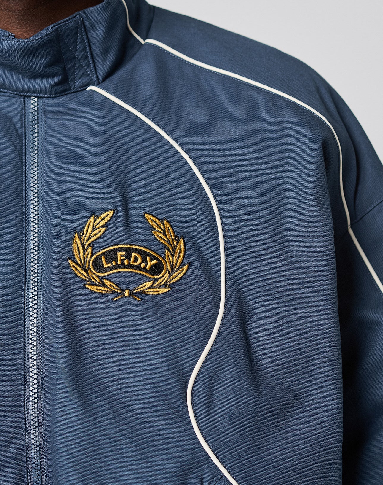 Close-up of the LFDY Canvas Jacket by LIVE FAST DIE YOUNG, featuring elastic cuffs, blue fabric, white piping, and an embroidered yellow laurel wreath with L.F.D.Y letters stitched in yellow on a black background.