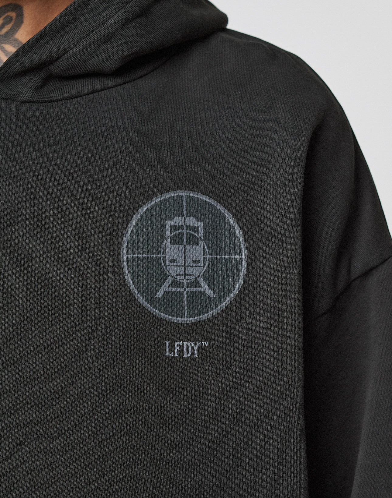 A close-up of the black LFDY Vandals Hooded by LIVE FAST DIE YOUNG shows a circular crosshair emblem with a stylized animal face and LFDY™ text. Made from 100% Baumwolle, part of the wearer’s neck and hood are visible.