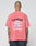 A person with a shaved head faces away, wearing the LIVE FAST DIE YOUNG Basic Evidence Tee in faded red. The back features repeated bold white and black LiveFast prints, a graphic, small white text below, and a subtle flag label on the hem.