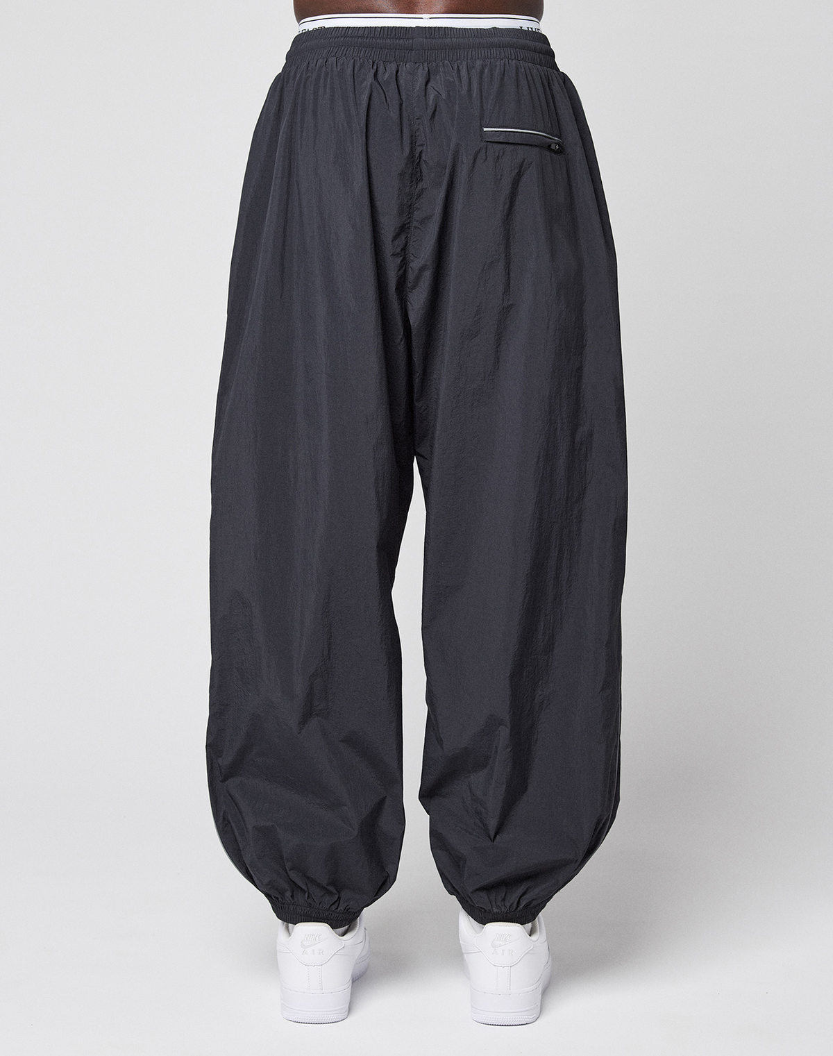 A person wears LIVE FAST DIE YOUNG's LFDY Performance Trackpants in black, featuring an oversize fit, 100% nylon material, elastic waistband and cuffs, and a zippered back pocket. White sneakers complete the look against a plain white background.