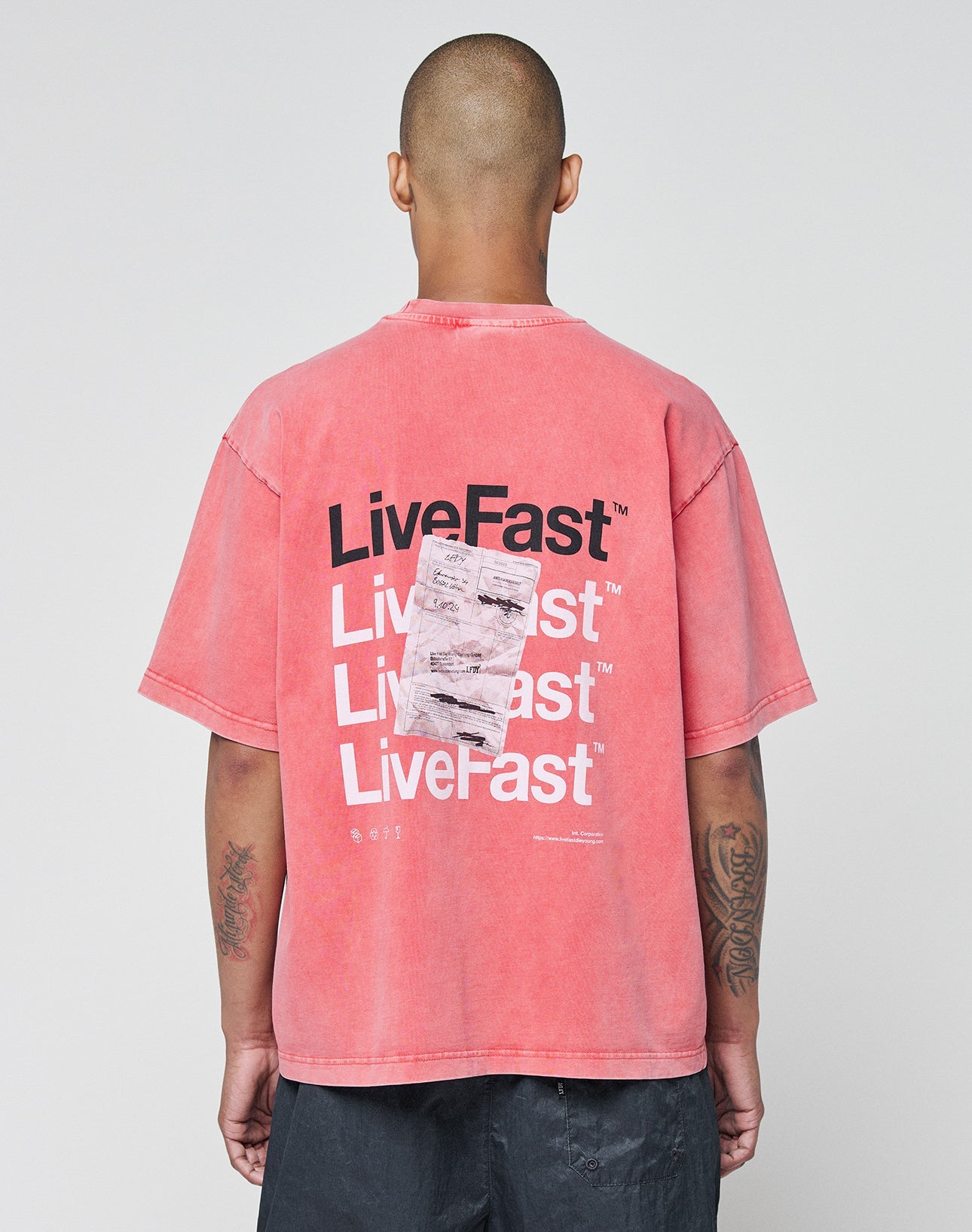 A person with a shaved head faces away, wearing the LIVE FAST DIE YOUNG Basic Evidence Tee in faded red. The back features repeated bold white and black LiveFast prints, a graphic, small white text below, and a subtle flag label on the hem.