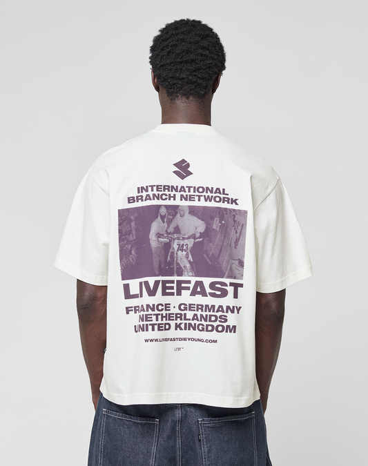 A person is seen from behind wearing the LFDY Grand Theft Tee by LIVE FAST DIE YOUNG, an oversize white 100% Baumwolle T-shirt with a vintage cycling photo and "INTERNATIONAL BRANCH NETWORK" listing France, Germany, Netherlands, and UK on the back.