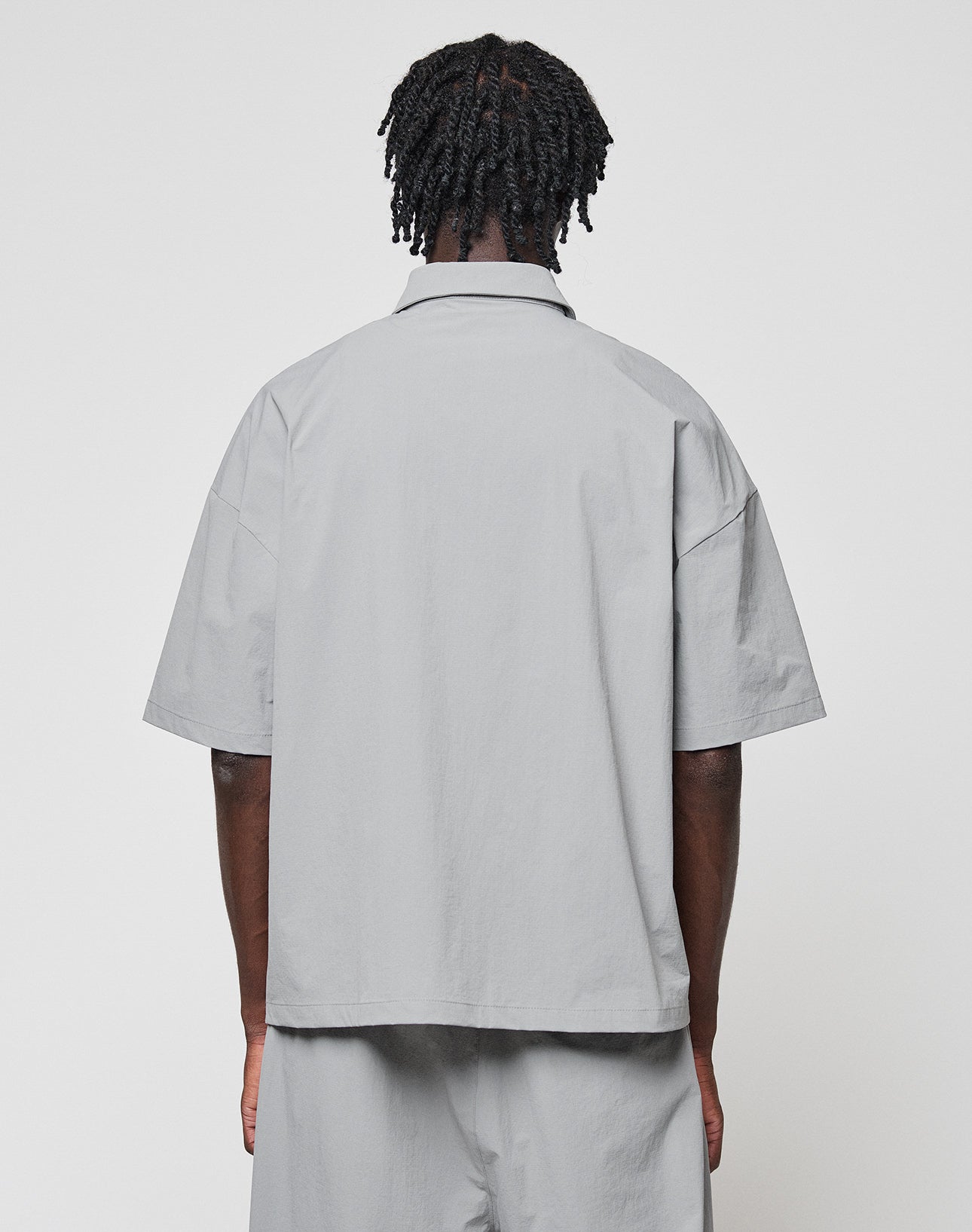 A person with short, twisted hair faces away from the camera, wearing an oversize fit, light gray LIVE FAST DIE YOUNG LFDY Ripstop Shirt and matching pants against a plain white background.