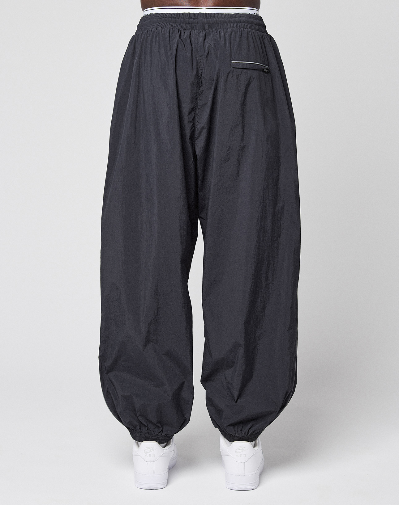 A person wears LIVE FAST DIE YOUNG's LFDY Performance Trackpants in black, featuring an oversize fit, 100% nylon material, elastic waistband and cuffs, and a zippered back pocket. White sneakers complete the look against a plain white background.