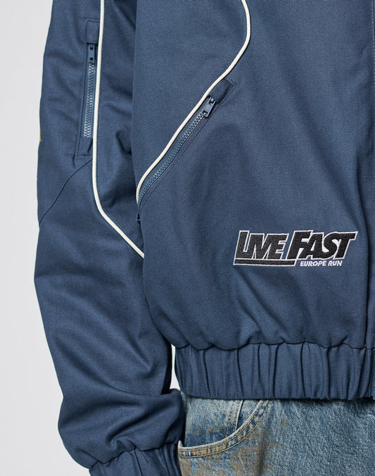 A close-up of a person wearing the blue LFDY Canvas Jacket by LIVE FAST DIE YOUNG, featuring white piping, EUROPE RUN logo, multicolor patches, elastic waistbands, zippered pockets, and paired with blue jeans.