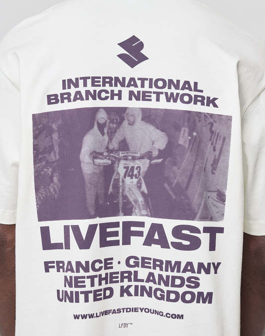 The LFDY Grand Theft Tee by LIVE FAST DIE YOUNG is a white oversize-fit shirt with purple front and back graphics of two people and a motocross bike, "International Branch Network LIVEFAST" text, country names, URL, and is 100% cotton.