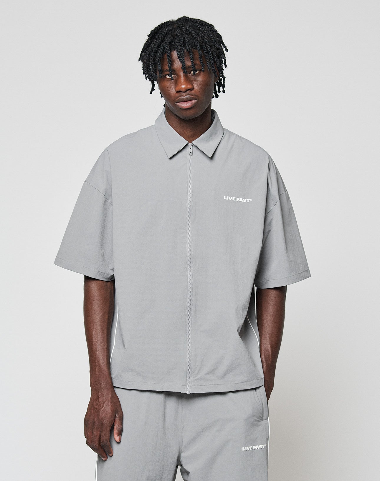 A man with short, twisted hair wears the LFDY Ripstop Shirt by LIVE FAST DIE YOUNG—an oversize fit, light gray zip-up shirt with a Kurzarm Hemdkragen and matching shorts featuring a bold LIVE FAST Silikonprint.