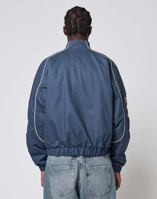 A person with braided hair is seen from behind, wearing the LFDY Canvas Jacket by LIVE FAST DIE YOUNG—blue with multilogo embroidery, white piping, elastic cuffs, and a high collar—paired with light blue jeans against a plain light background.