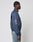A person stands in profile against a plain background, wearing the LFDY Canvas Jacket by LIVE FAST DIE YOUNG with white piping, multicolored logo embroidery on the sleeve, and light blue jeans.