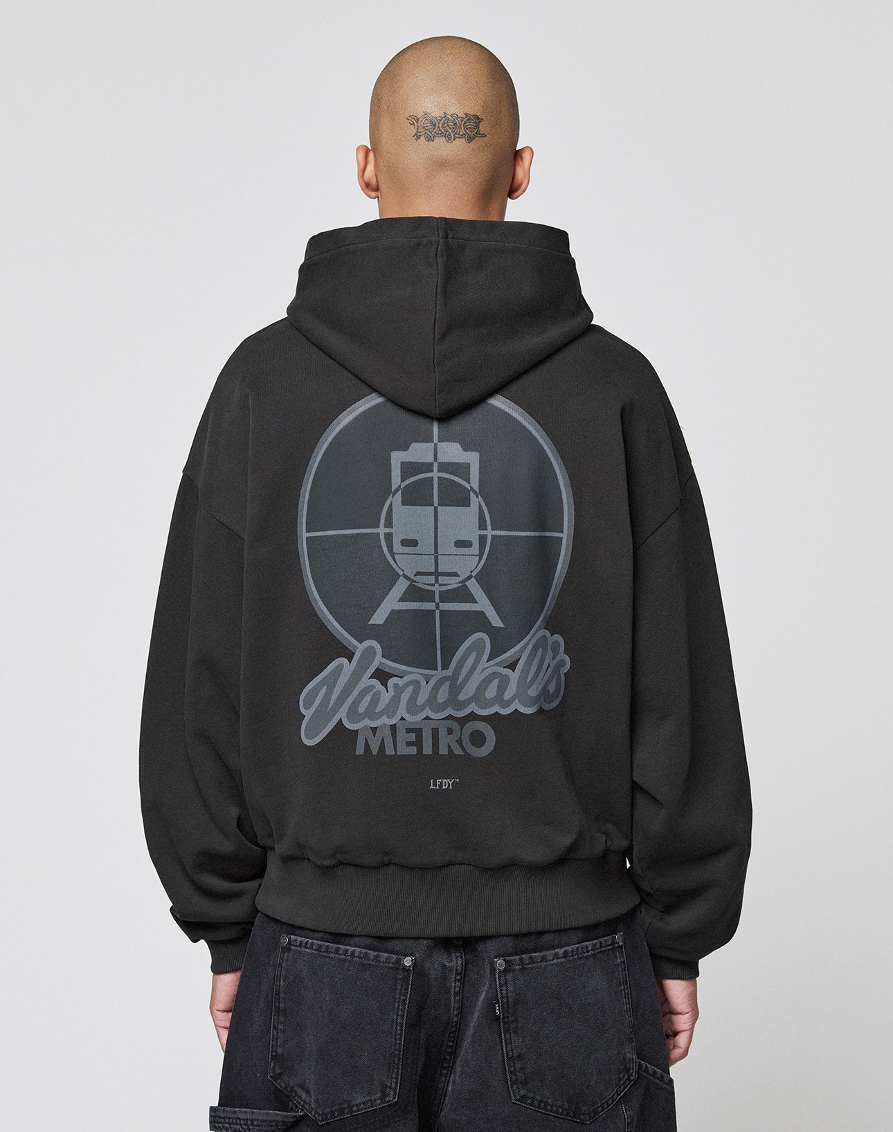 A person with a shaved head and neck tattoo, facing away, wears the LFDY Vandals Hooded by LIVE FAST DIE YOUNG—an oversized black 100% cotton hoodie featuring a subway train in crosshairs and "Vandals METRO" on the back.