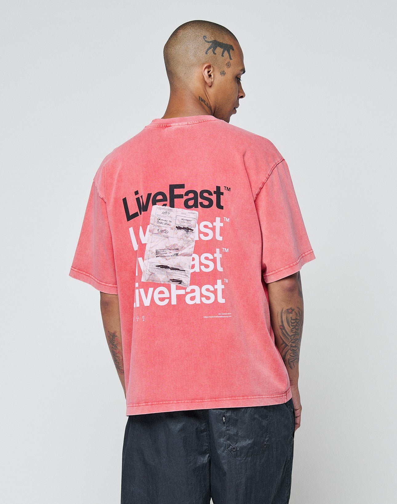 A tattooed, shaved-head person wears the LIVE FAST DIE YOUNG Basic Evidence Tee—an oversize, faded red 100% Baumwolle shirt with “Live Fast” in bold white and black plus a white graphic on the back and a subtle flag-label at the hem.