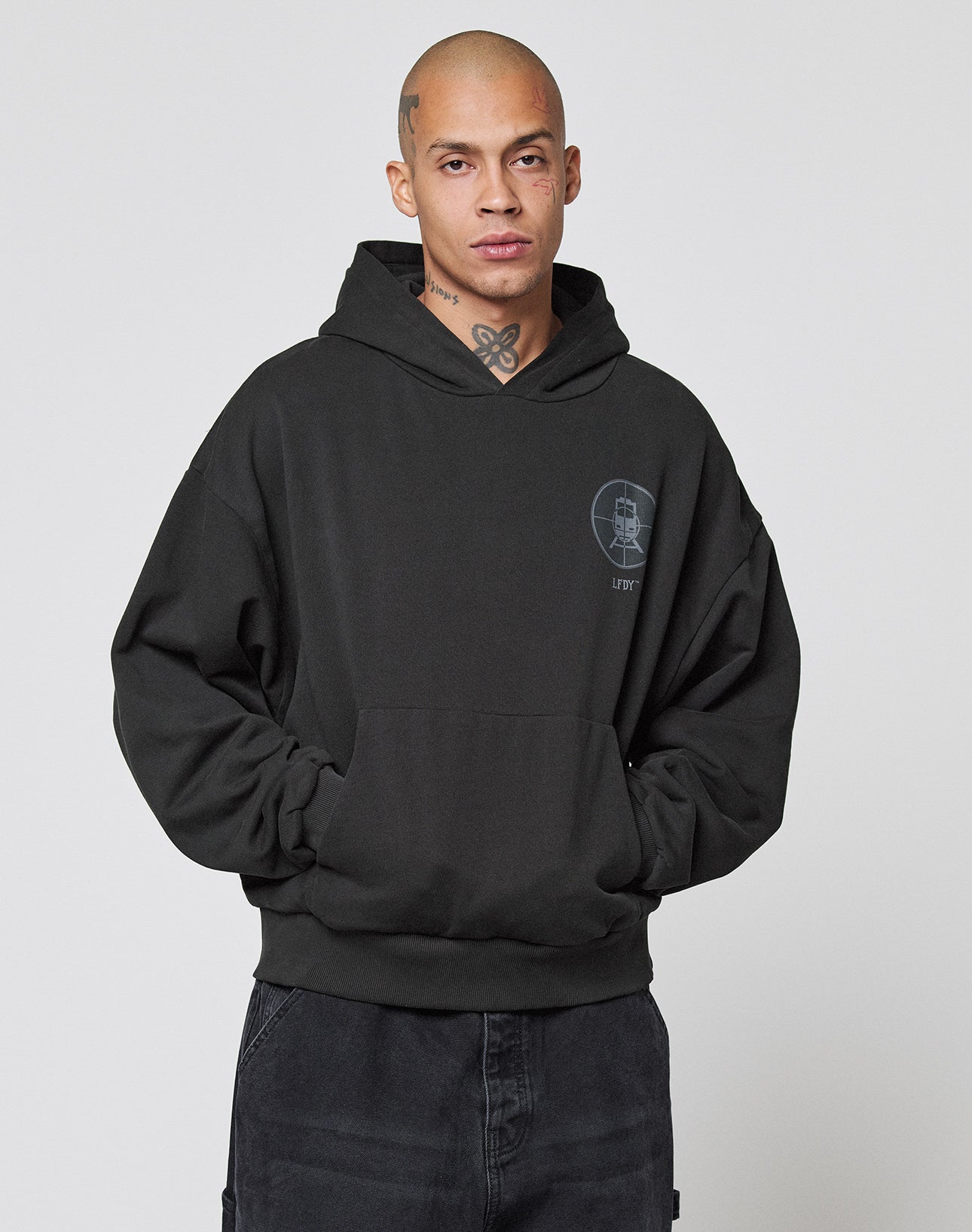 A person with a shaved head and neck tattoos wears the LFDY Vandals Hooded by LIVE FAST DIE YOUNG, featuring a small chest graphic, standing with hands in pockets against a plain light background.