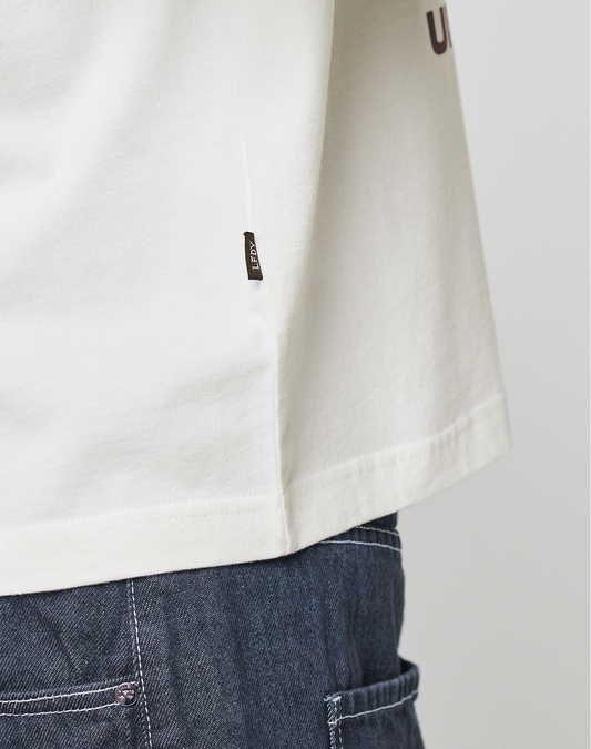 A close-up of a person wearing the LFDY Grand Theft Tee by LIVE FAST DIE YOUNG, featuring an oversize fit with a white 100% Baumwolle fabric, a small black side tag, and paired with blue denim jeans. Focus is on the t-shirt hem and jeans’ waist.