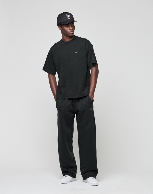 A person poses against a plain background wearing the oversized black 'Basic Evidence Tee' by LIVE FAST DIE YOUNG, paired with black pants, white sneakers, and a black baseball cap. Hands in pockets, they gaze to the side.