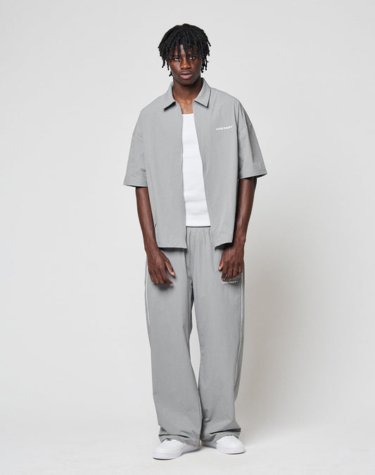 A man stands against a plain white background wearing a loose-fitting light gray short-sleeve shirt over a white t-shirt, LIVE FAST DIE YOUNG LFDY Ripstop Trackpants in gray with an elastic waistband, and white sneakers. He faces forward neutrally.