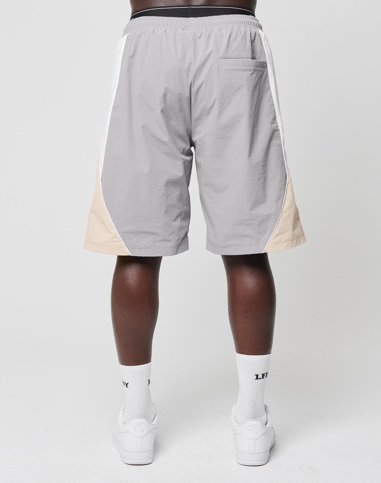 A person stands with their back to the camera against a plain background, wearing LIVE FAST DIE YOUNG Basic Nylon Trackshorts in light gray with beige side panels, paired with white socks and sneakers.