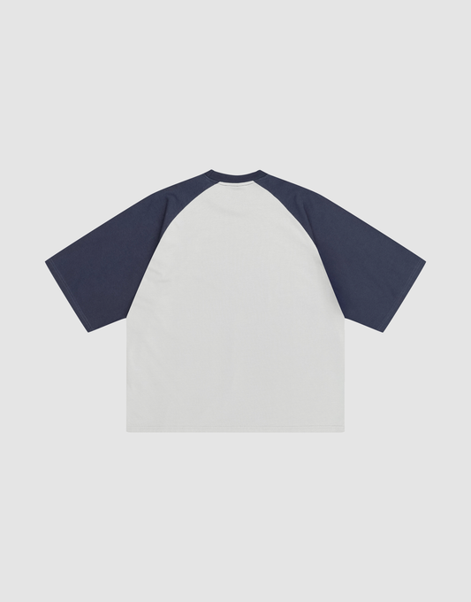 The LIVE FAST DIE YOUNG Live Fast Raglan Tee features a white body with dark navy raglan sleeves, crafted from 100% cotton. Displayed flat on a white background, the image shows the back view of this short-sleeve tee.