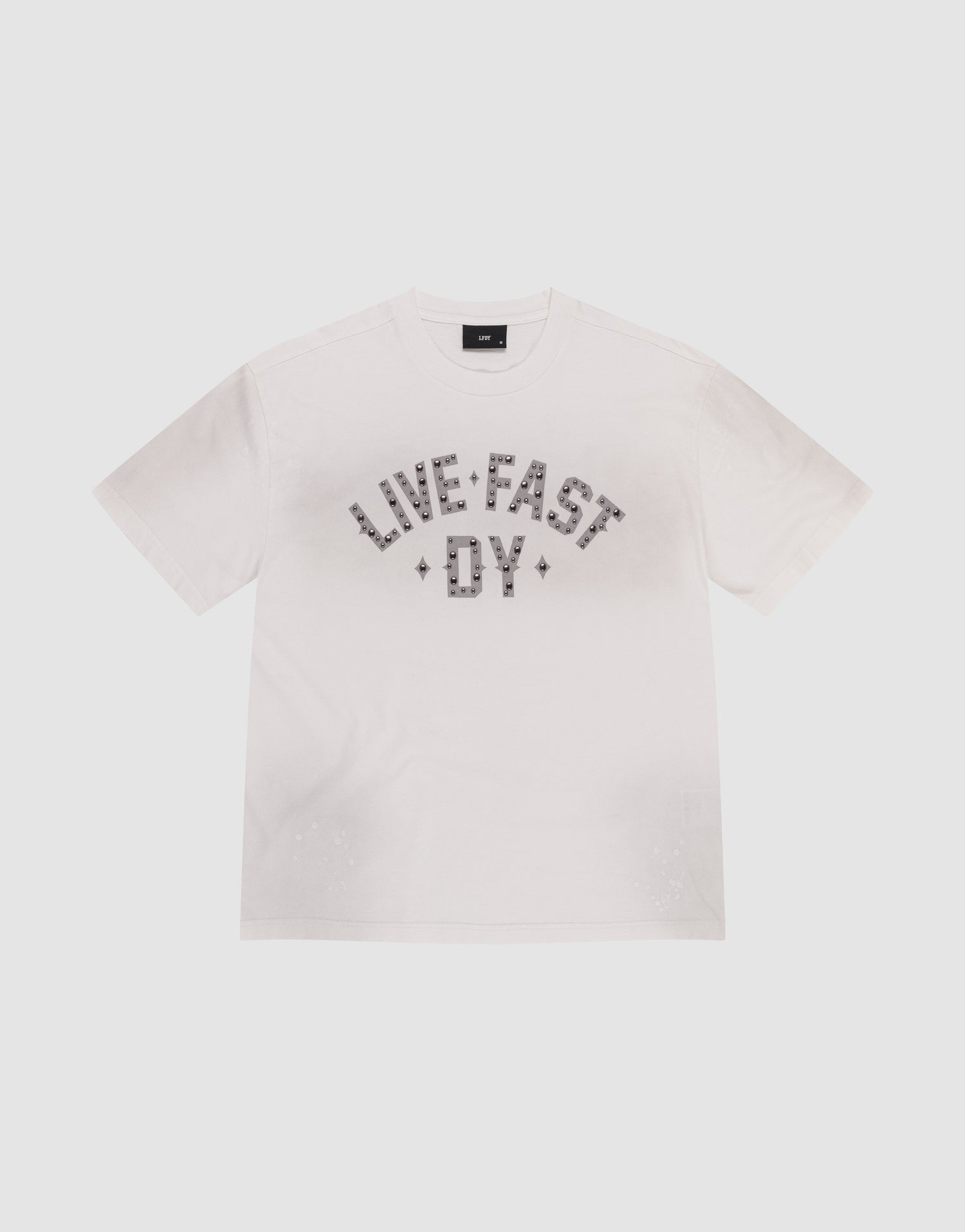 The Live Fast Rivet Tee by LIVE FAST DIE YOUNG is a white short-sleeve t-shirt featuring "LIVE FAST DIE YOUNG" in bold, patterned letters across the chest and subtle distressing for a worn-in edge. Displayed flat on a light background. | plain