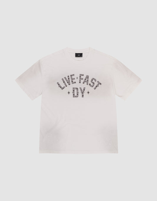 The Live Fast Rivet Tee by LIVE FAST DIE YOUNG is a white short-sleeve t-shirt featuring "LIVE FAST DIE YOUNG" in bold, patterned letters across the chest and subtle distressing for a worn-in edge. Displayed flat on a light background.