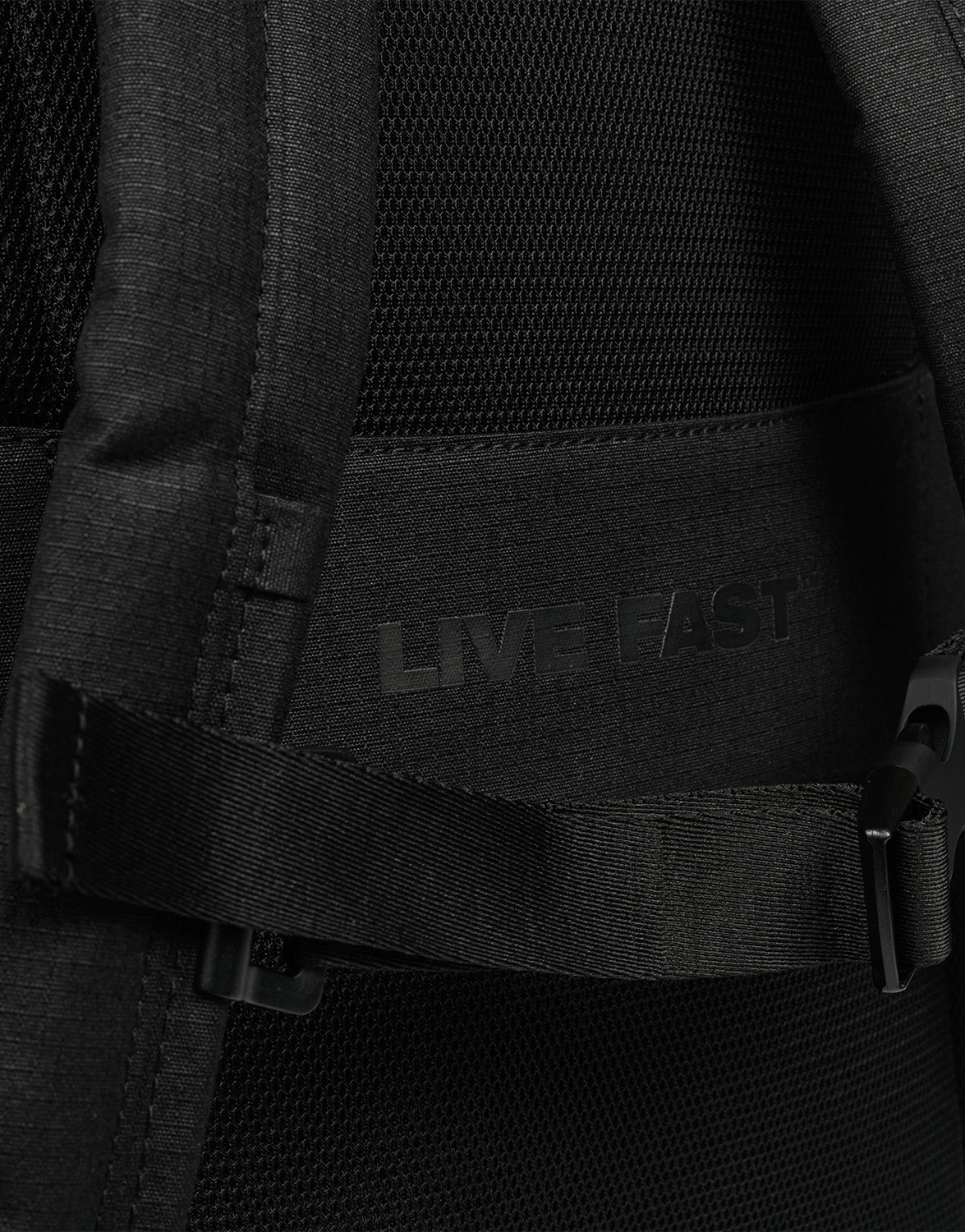 Close-up of the black LFDY Backpack (19L) by LIVE FAST DIE YOUNG, with "LIVE FAST" subtly printed on the fabric, partially covered by a strap and plastic buckle, and featuring adjustable shoulder straps for comfort.
