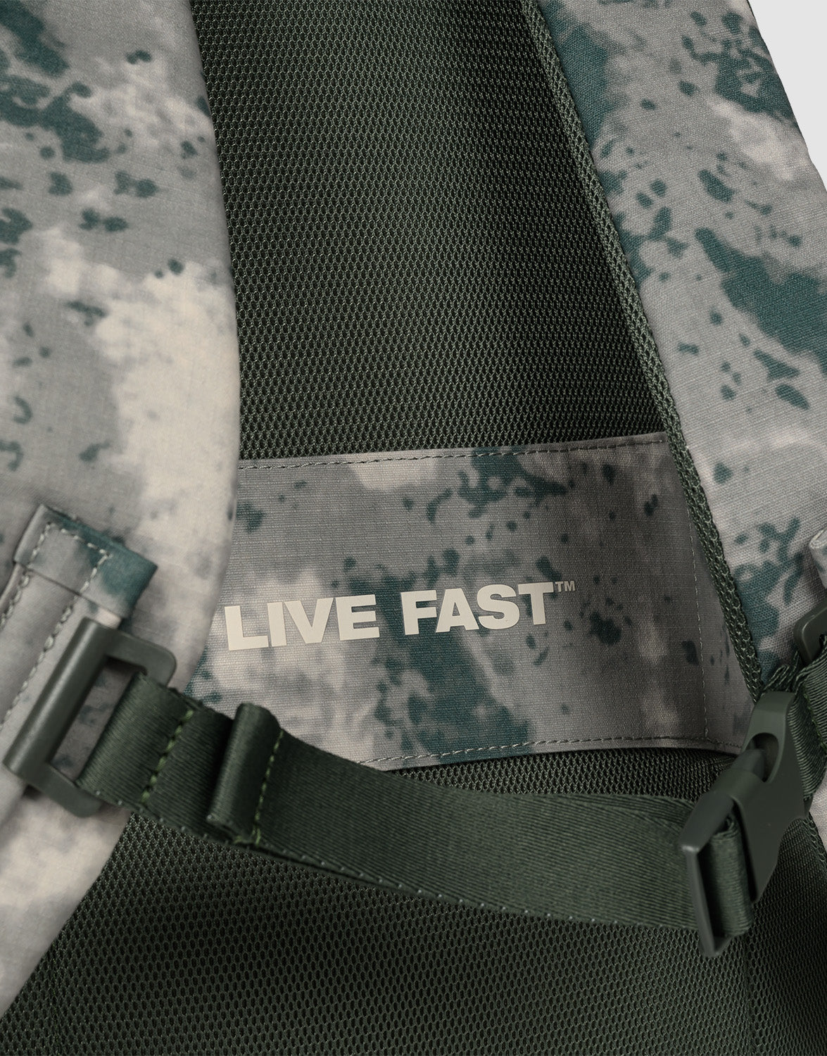 Close-up of the LFDY Travel Backpack by LIVE FAST DIE YOUNG, a green and gray camo 25L rucksack featuring a mesh panel, adjustable shoulder straps, and an inner label reading "LIVE FAST" in bold white letters.