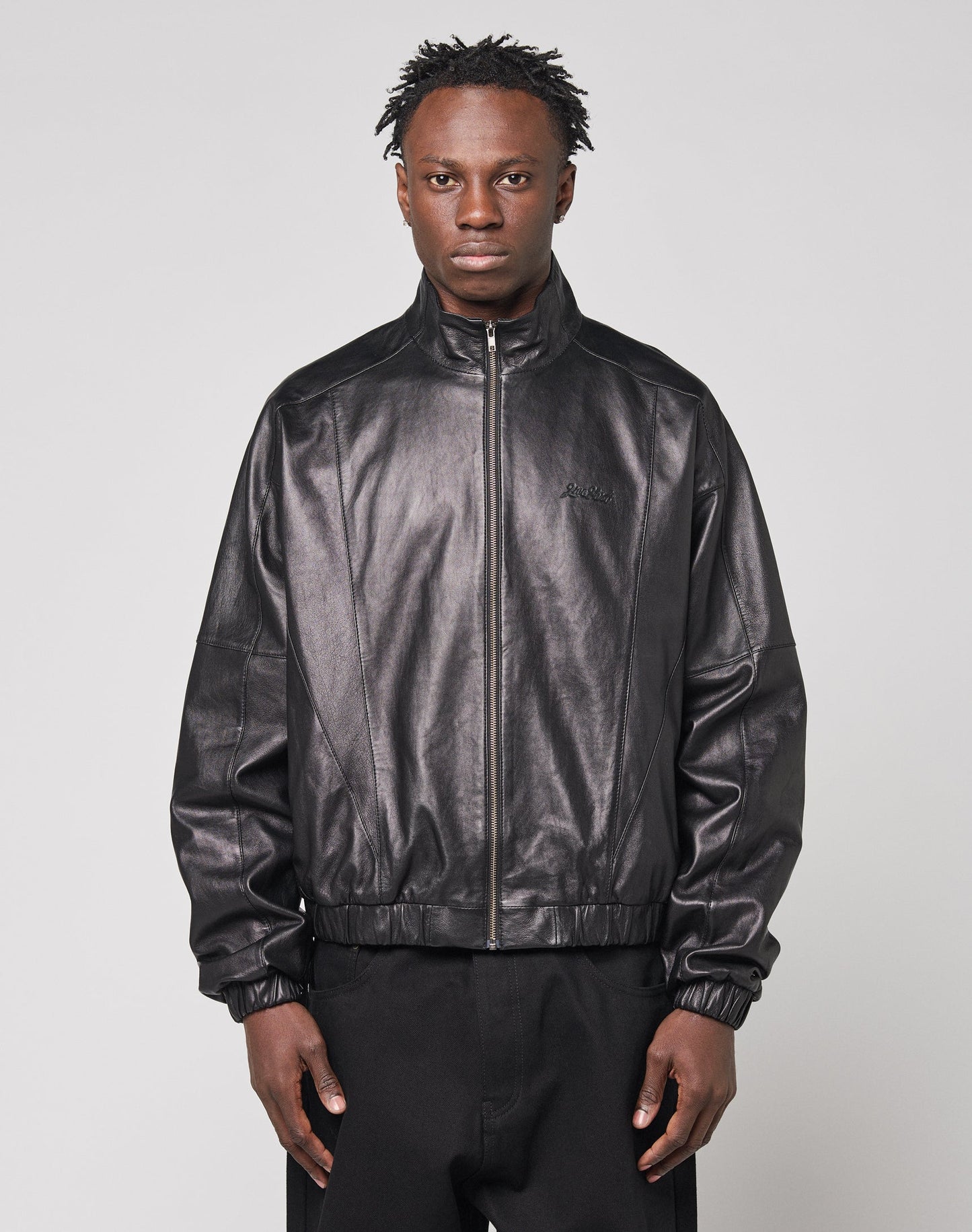 A person stands facing forward, wearing the LIVE FAST DIE YOUNG Live Fast Leather Trackjacket in black with Stehkragen and embossed logo, paired with black pants, against a plain light gray background.