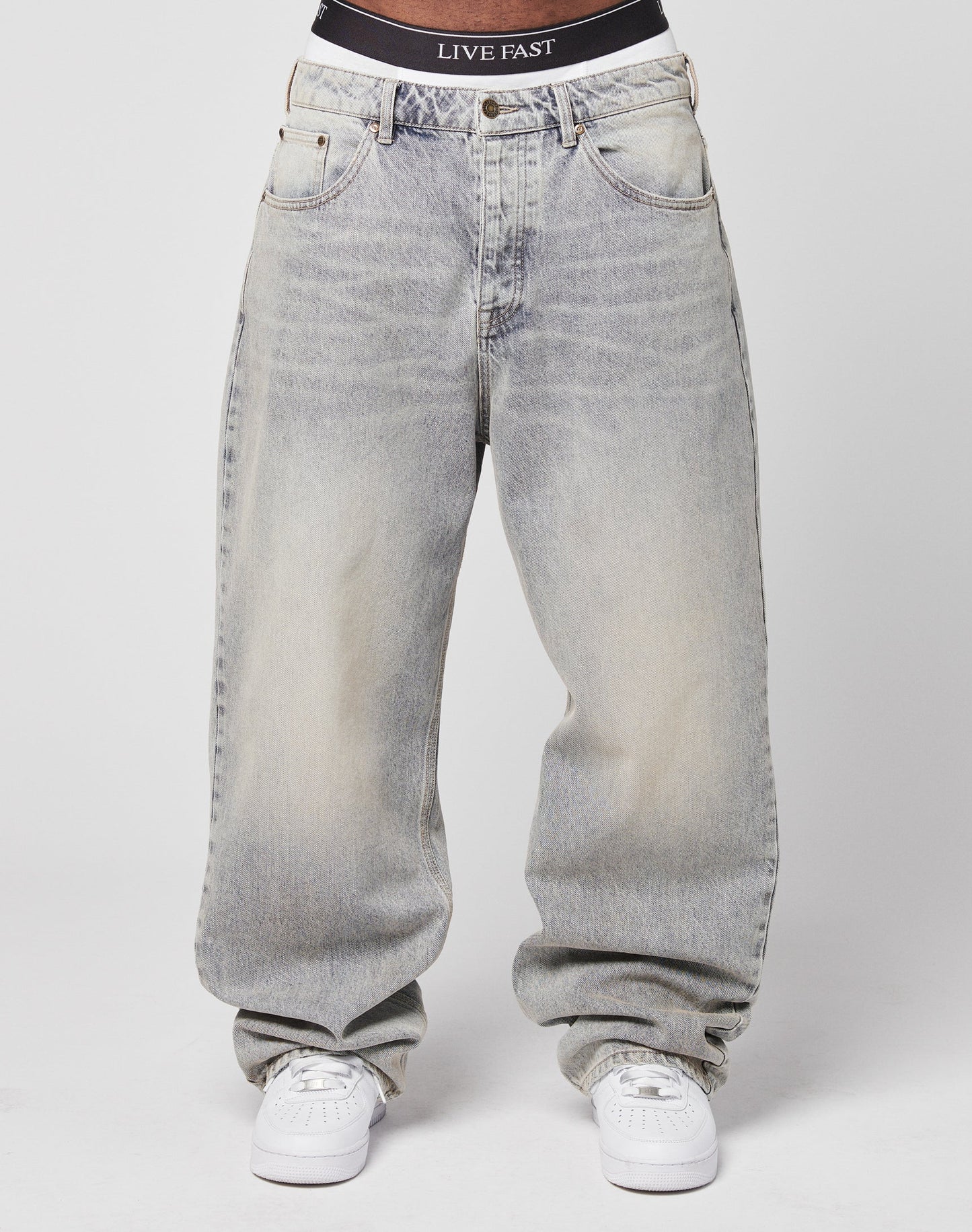 A person wears the LFDY Baggy Denim—light-wash, baggy fit with a wide straight leg—paired with white sneakers. A black waistband featuring the LIVE FAST slogan is visible above the jeans. The background is plain white.