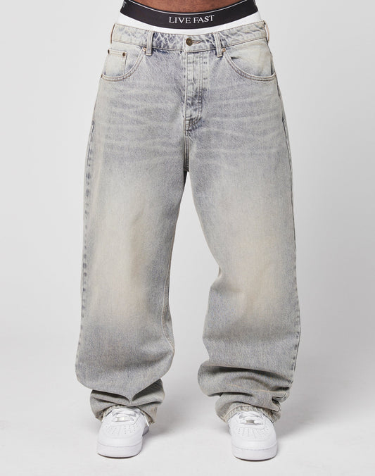 Someone is wearing LFDY Baggy Denim by LIVE FAST DIE YOUNG—light-wash, 100% Baumwolle jeans with a wide straight leg—paired with white sneakers and black LIVE FAST waistband underwear. The background is plain white.
