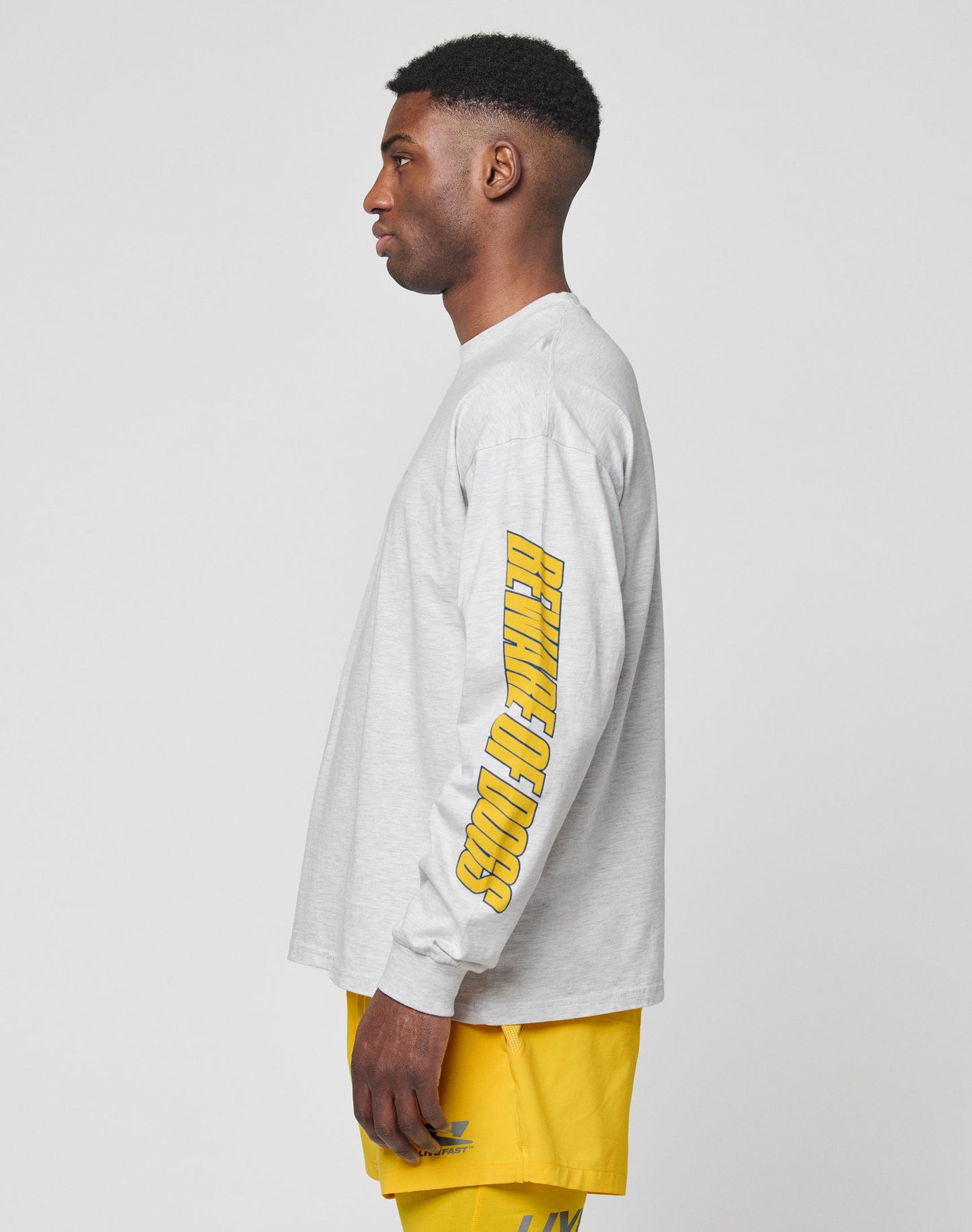 A man in a light gray LFDY Split Sphere Longsleeve by LIVE FAST DIE YOUNG, featuring “REVIVAL OF GOODS” in yellow on the sleeve, and yellow shorts stands in profile against a plain light background.