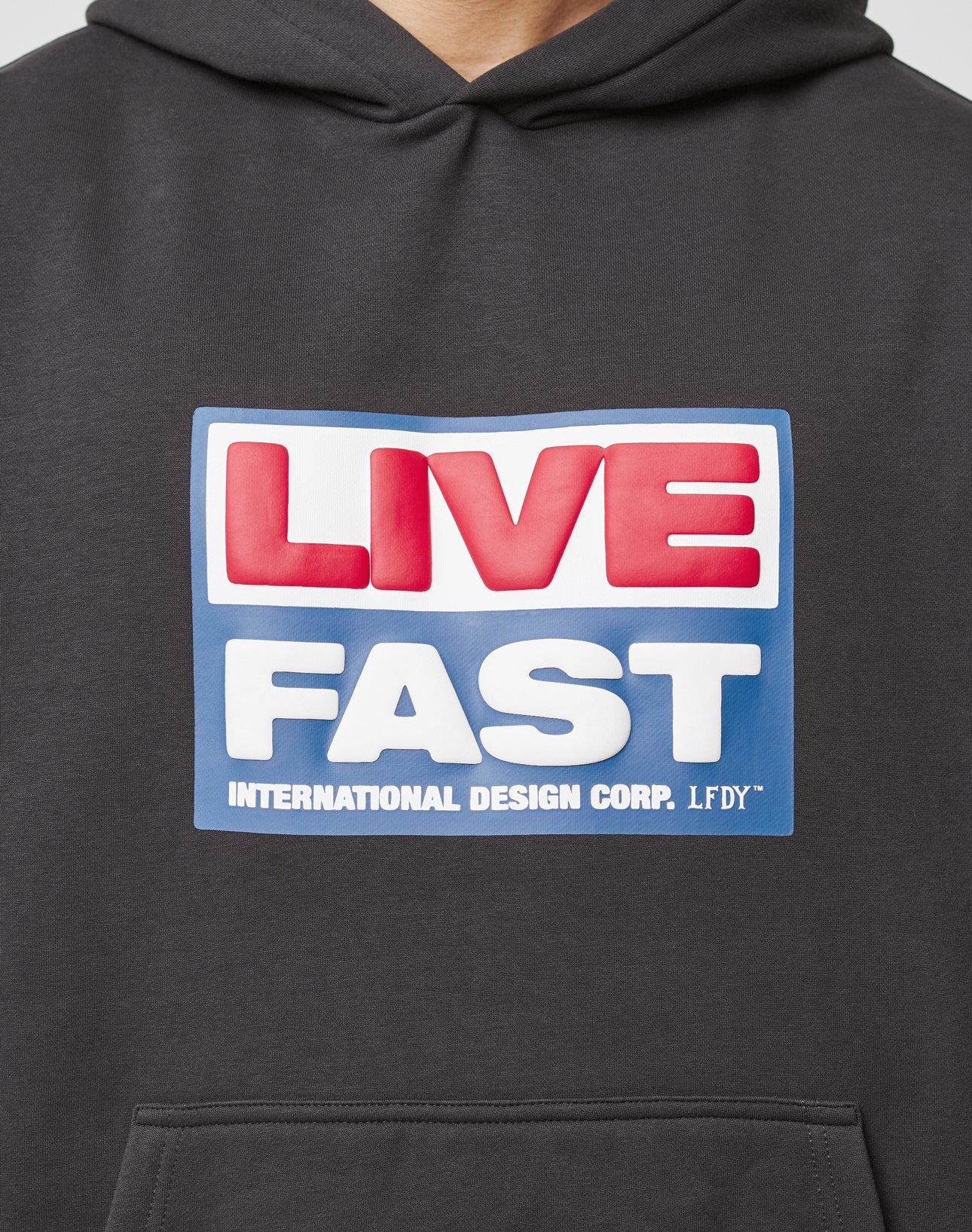 The Live Fast Box Hooded by LIVE FAST DIE YOUNG is a black, oversized hoodie with a bold puffer print: LIVE in red on a white and blue background above FAST in white on blue; INTERNATIONAL DESIGN CORP. LFDY below and a flag-label accent.