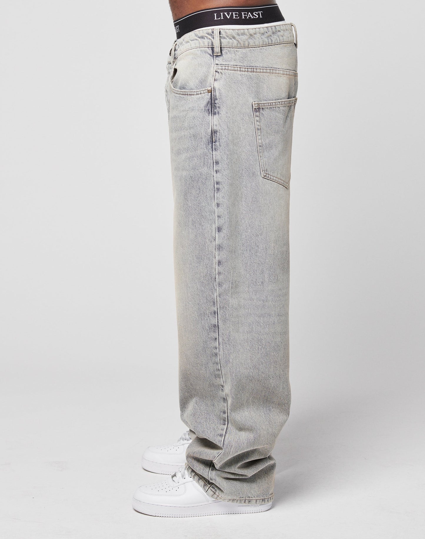 A person stands sideways in light-wash LFDY Baggy Denim by LIVE FAST DIE YOUNG, featuring a straight wide leg and subtle flag label, styled with white sneakers and black underwear showing LIVE FAST above the waistband against a plain white backdrop.