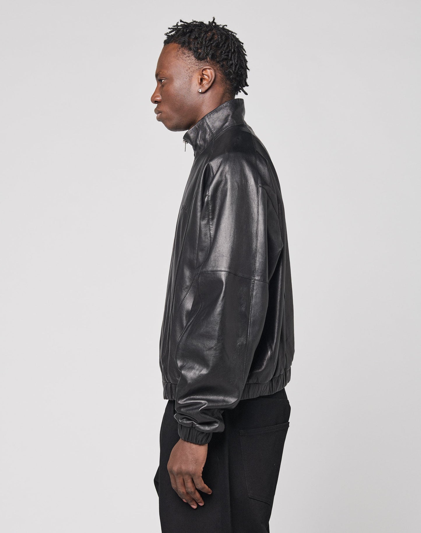 A person with short, textured hair is shown in profile against a plain background, wearing the LIVE FAST DIE YOUNG Live Fast Leather Trackjacket in black and black pants.