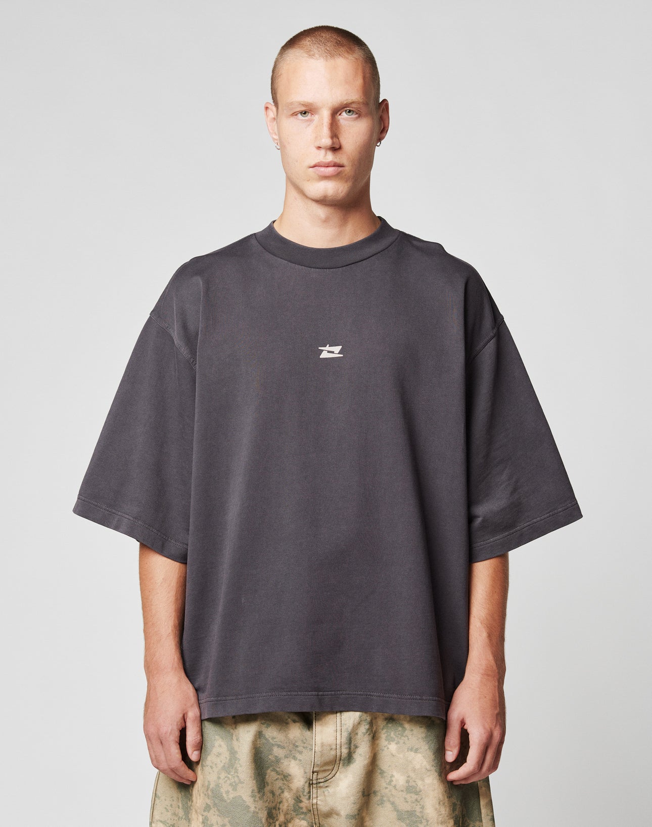 A person with short hair wears the LIVE FAST DIE YOUNG Live Fast Cropped Oversized Tee in dark gray with a white center logo, paired with light camo pants, standing against a plain light gray background.