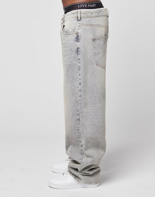 A person stands in profile against a white background wearing LIVE FAST DIE YOUNG’s LFDY Baggy Denim jeans, white sneakers, and black boxer briefs with LIVE FAST on the waistband.