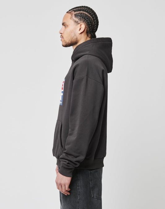 A man with braided hair stands in profile against a light gray background, wearing the LIVE FAST DIE YOUNG Live Fast Box Hooded sweatshirt in black and faded black jeans.