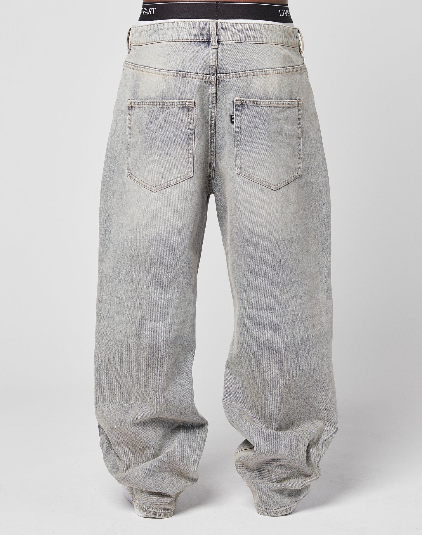 Seen from the back, a person wears LIVE FAST DIE YOUNG’s LFDY Baggy Denim—loose, faded gray jeans with a wide straight leg, two back pockets, a visible waistband, subtle flag label—all against a plain light background.