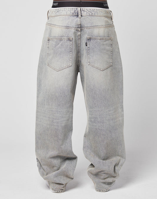 Seen from the back, a person wears LIVE FAST DIE YOUNG’s LFDY Baggy Denim—loose, faded gray jeans with a wide straight leg, two back pockets, a visible waistband, subtle flag label—all against a plain light background.