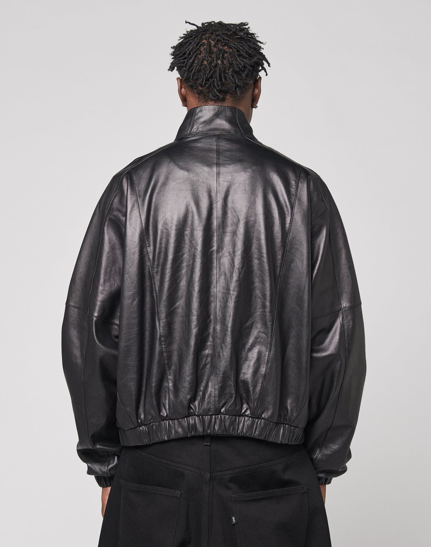 A person with short, twisted hair is shown from behind wearing the LIVE FAST DIE YOUNG Live Fast Leather Trackjacket with Stehkragen and black pants, standing against a plain light gray background.