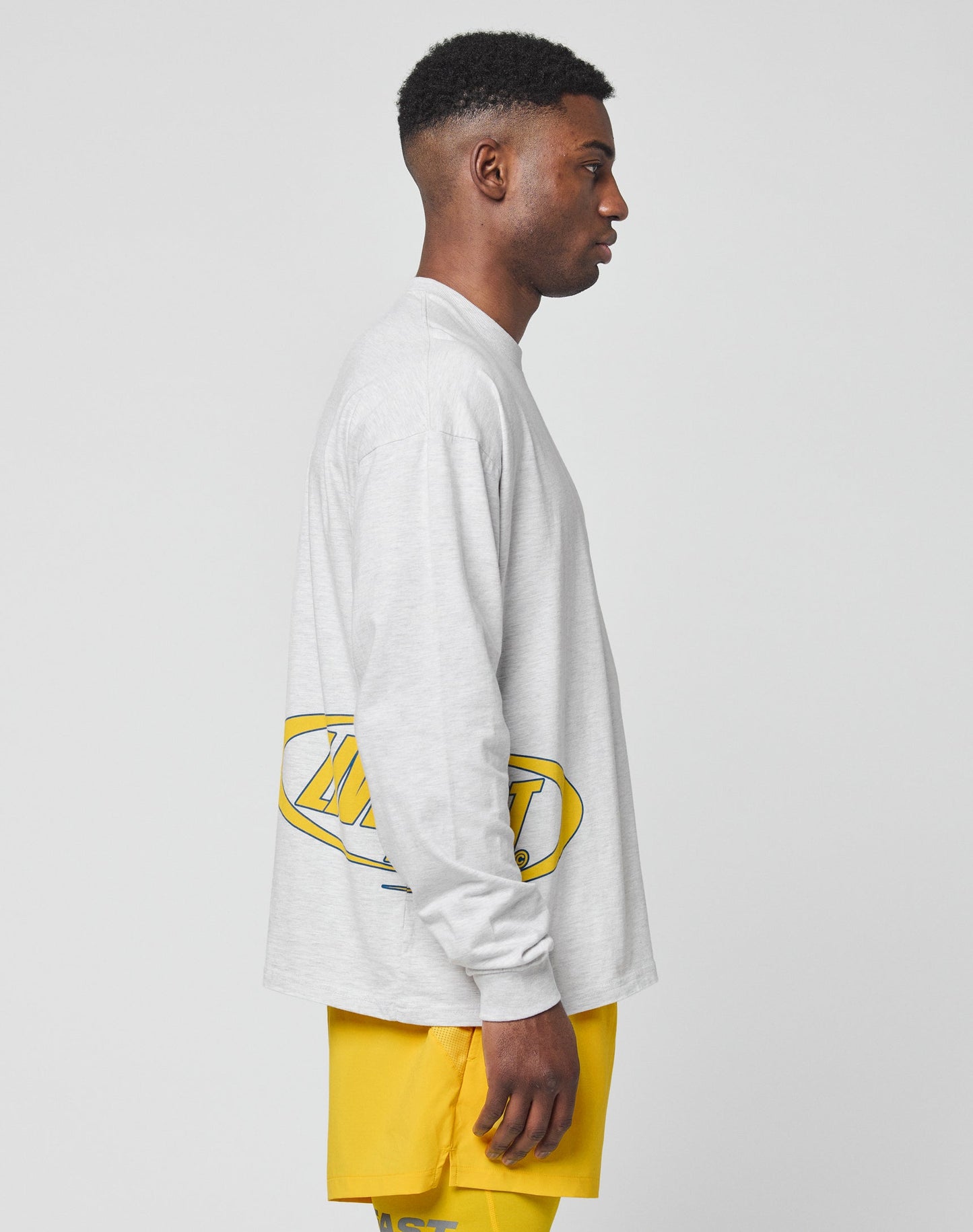 A man stands in profile against a plain background, wearing the LIVE FAST DIE YOUNG LFDY Split Sphere Longsleeve in light gray with a yellow and black graphic on the back, paired with bright yellow shorts.