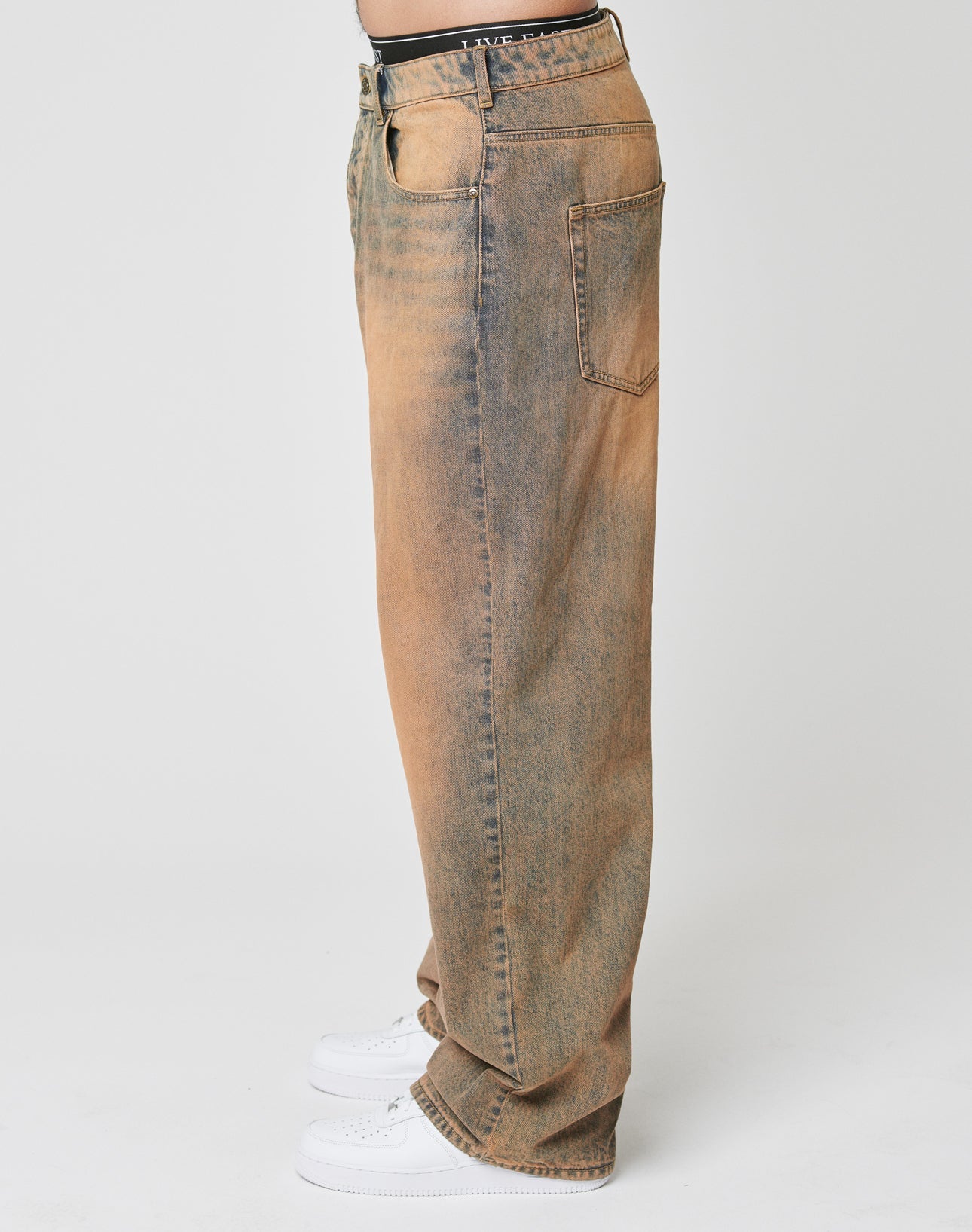 A person in profile wears LFDY Baggy Denim jeans by LIVE FAST DIE YOUNG—loose-fitting, faded brown and blue, with a baggy fit and wide straight leg. Made from 100% cotton, featuring front and back pockets, paired with white sneakers on a plain white background.