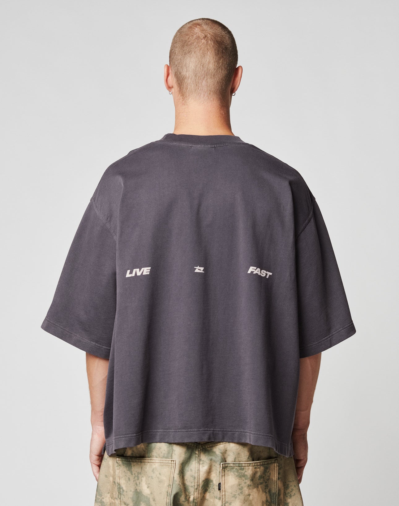 A person with a buzz cut, seen from behind, wears the LIVE FAST DIE YOUNG Live Fast Cropped Oversized Tee in dark gray with a bold backprint and star detail, paired with light green and beige tie-dye pants.
