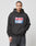 A man with braided hair wears the LIVE FAST DIE YOUNG Live Fast Box Hooded, an oversized black hoodie featuring a large front pocket and bold red, white, and blue puffer print, standing against a plain white background.